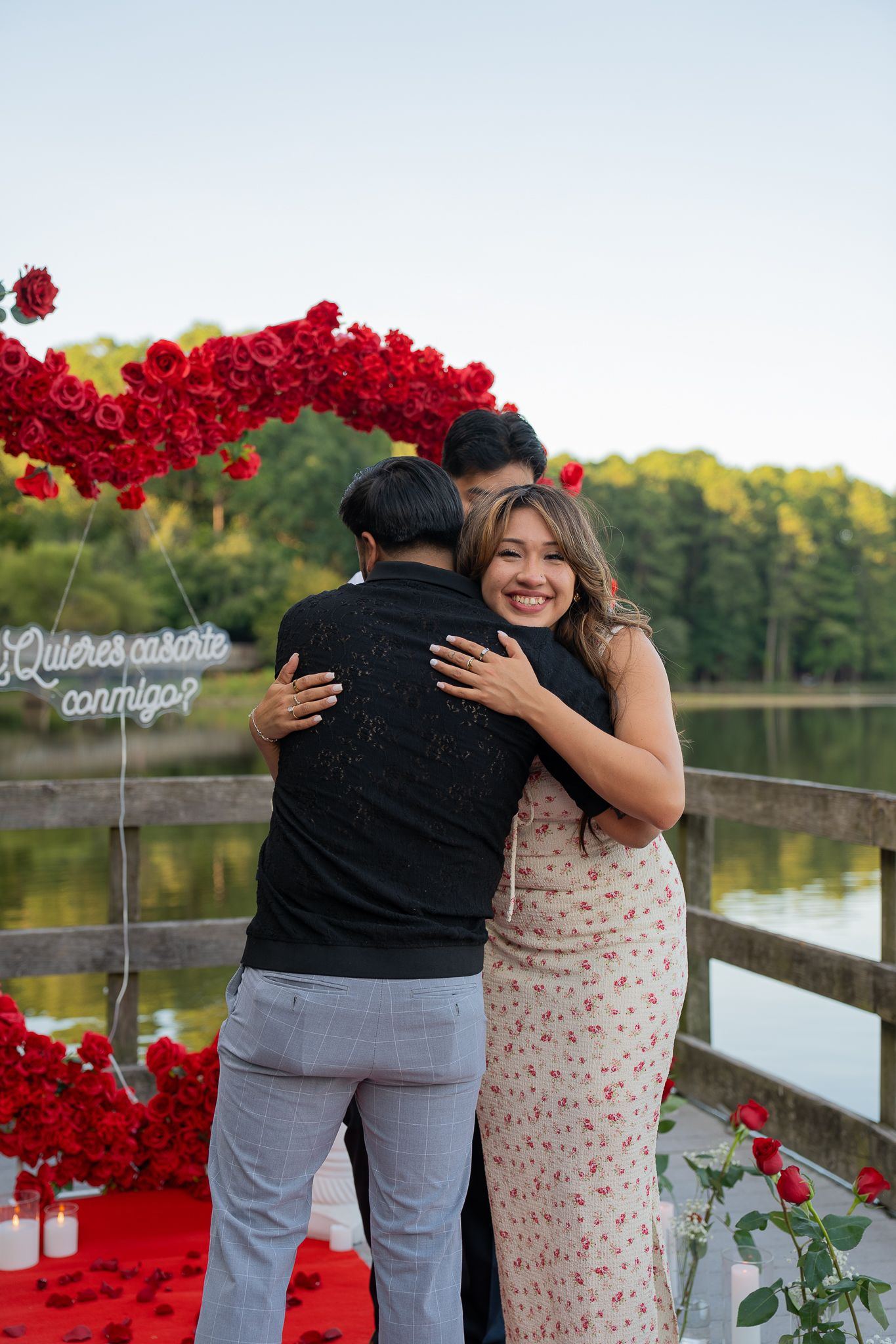Pareja abrazándose junto a un lago, con un corazón de rosas rojas al fondo