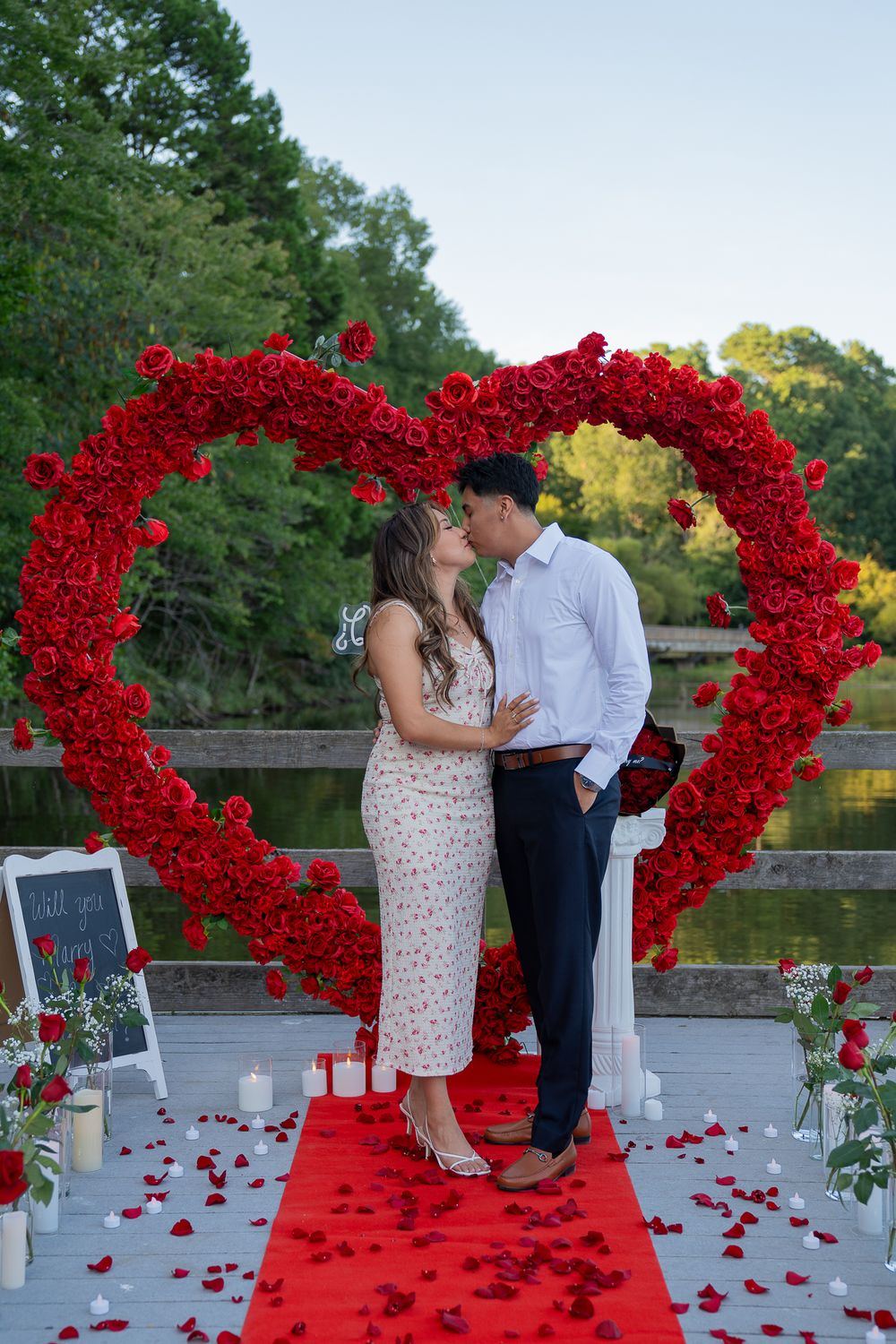 Pareja frente a un gran corazón de rosas rojas sobre una alfombra roja junto al lago