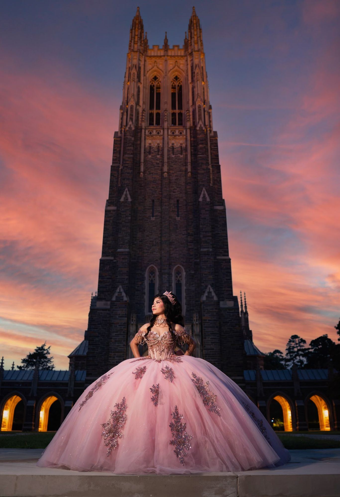 Quinceañera con vestido rosa bordado y corona, de pie frente a la torre de una capilla gótica bajo un cielo rosado al atardecer