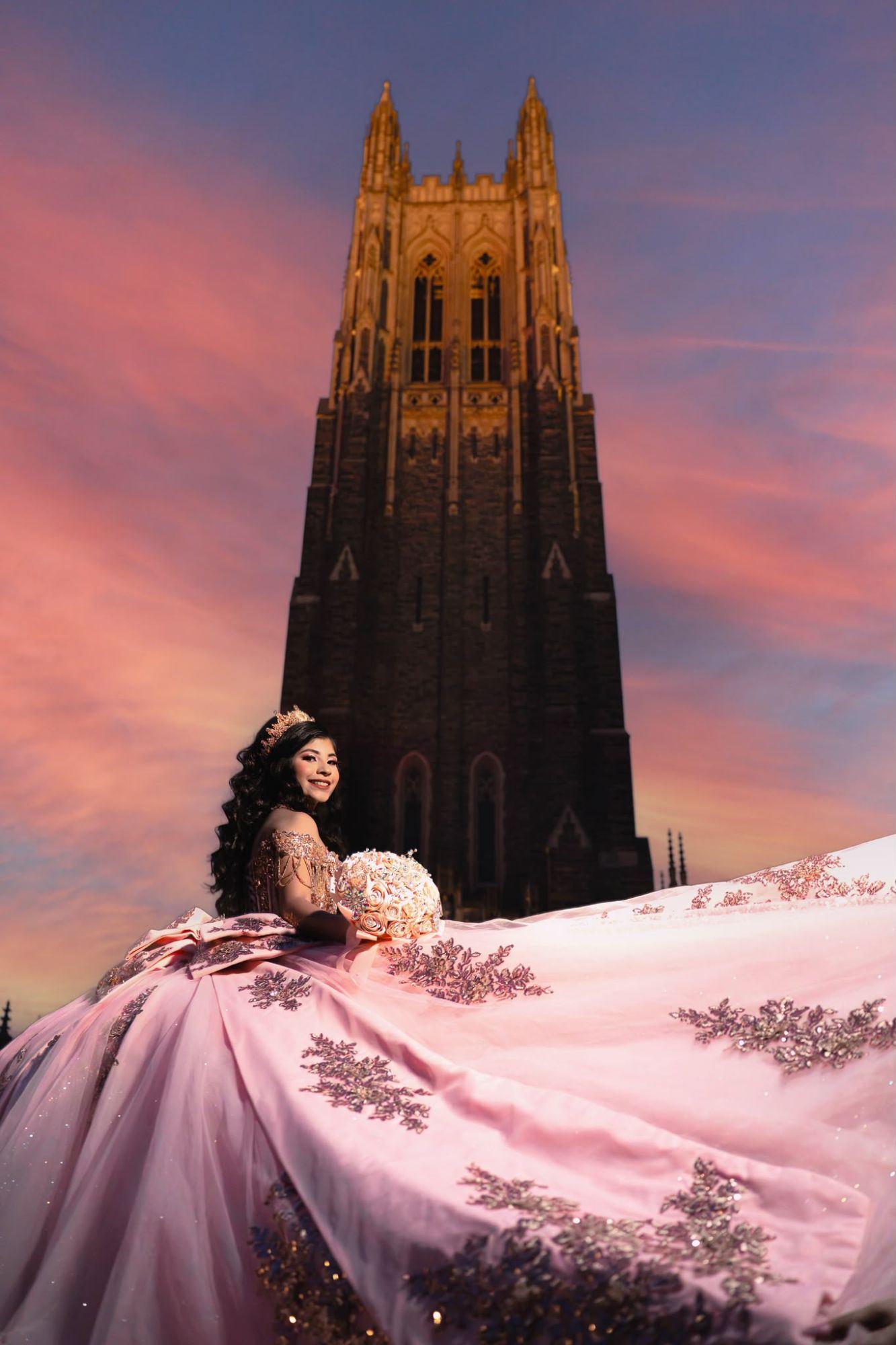 Quinceañera sonriente con un ramo, sentada sobre la amplia cola de su vestido rosa frente a la torre de una capilla al atardecer