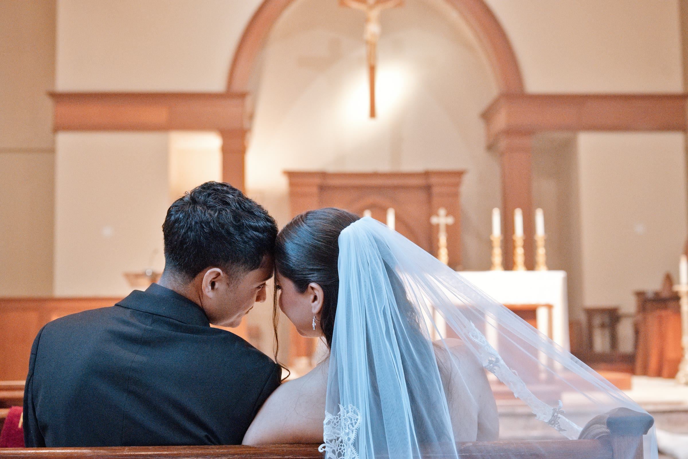 Novios sentados en la primera fila de la iglesia, juntando la frente frente al altar